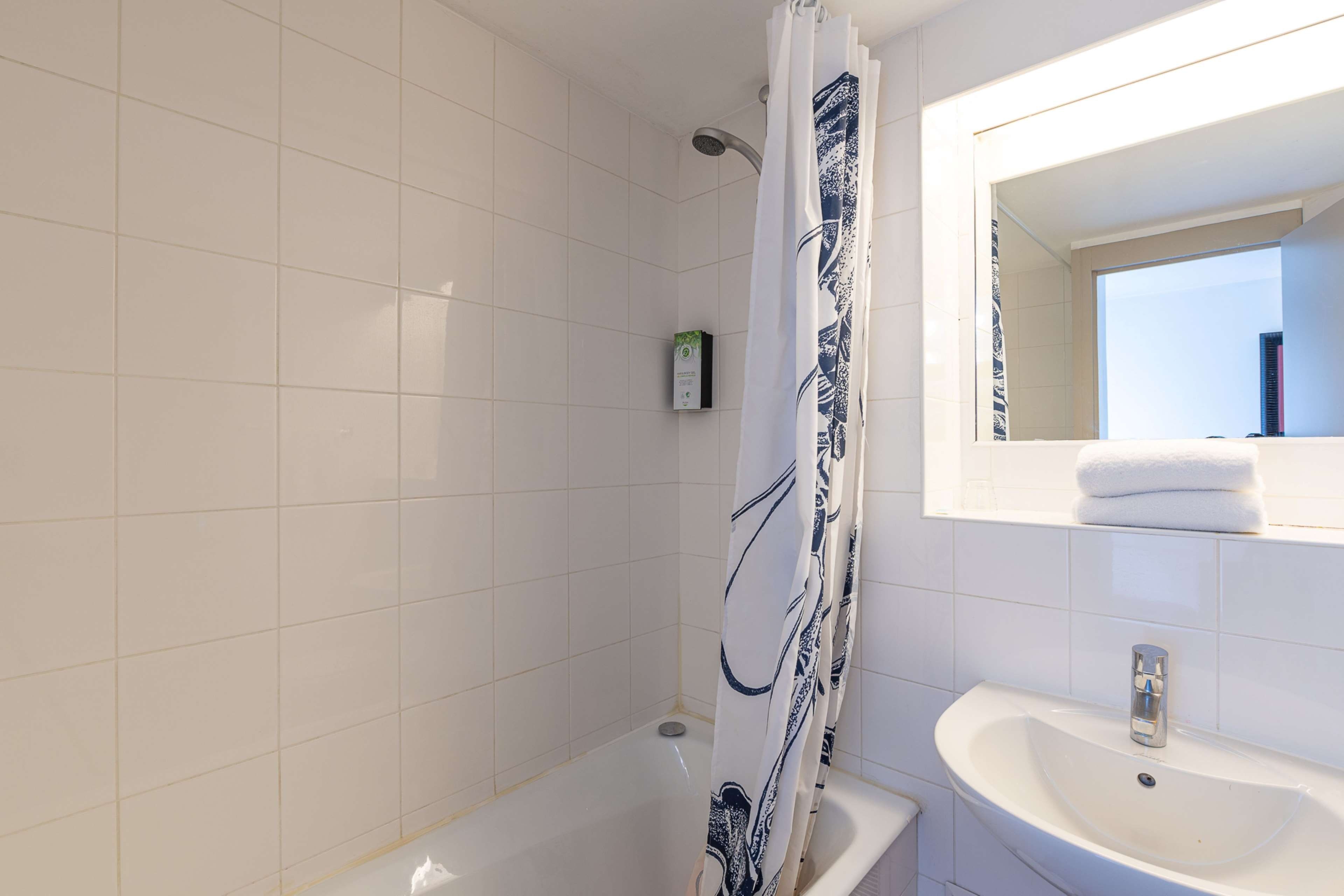 A bathroom with white tiles, a shower curtain with a blue pattern, a sink with a mirror above it, and a small plant on the wall.