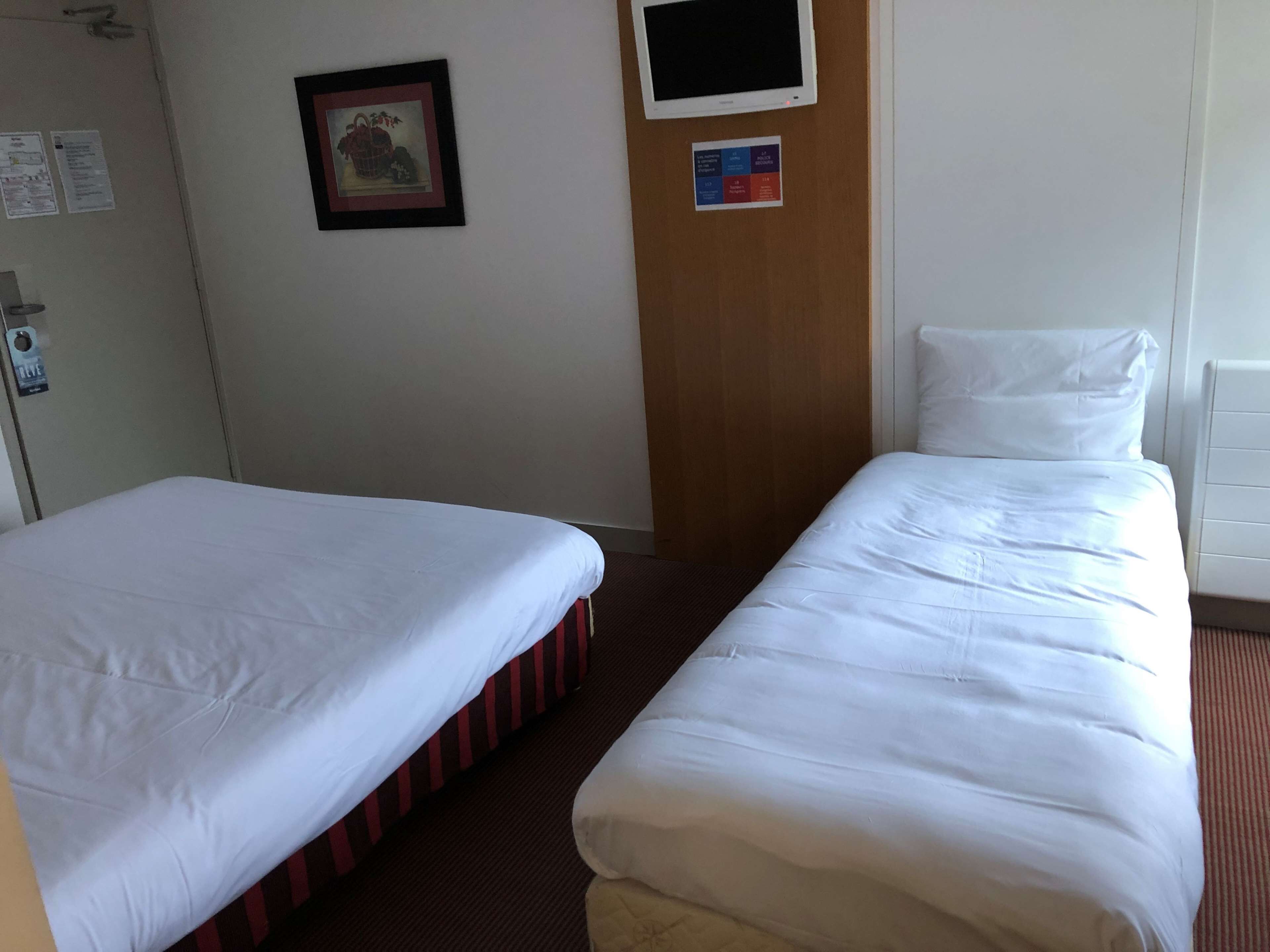 Une chambre d’hôtel avec deux lits simples, une porte en bois, un tableau encadré au mur et un petit écran numérique.