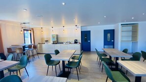Een moderne eetruimte met meerdere tafels en stoelen, een keukenhoek met koffieapparaat, en een blauwe deur.