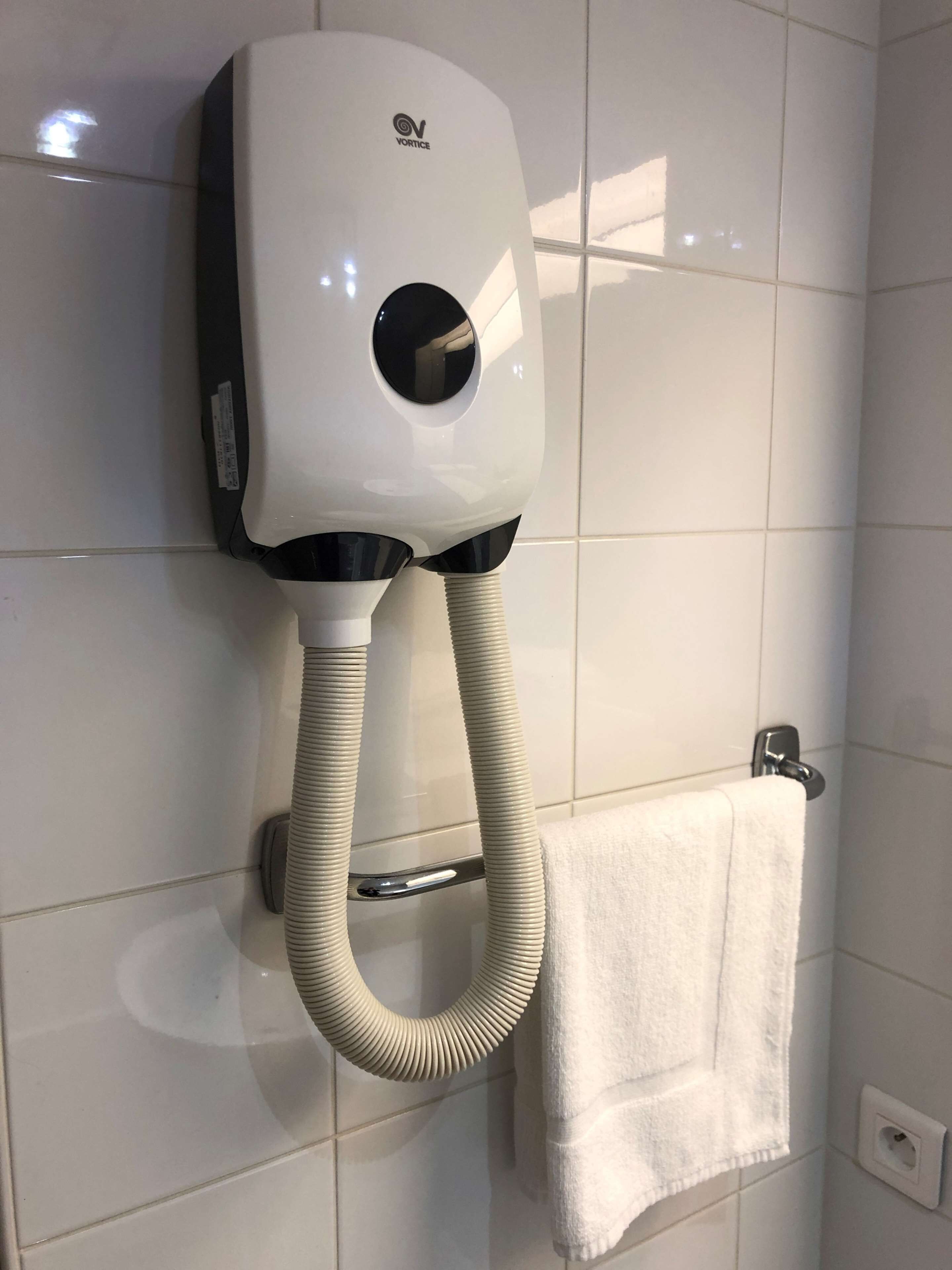 Een aan de muur bevestigde haardroger met flexibele slang, een handdoekhouder en een witte handdoek eraan.