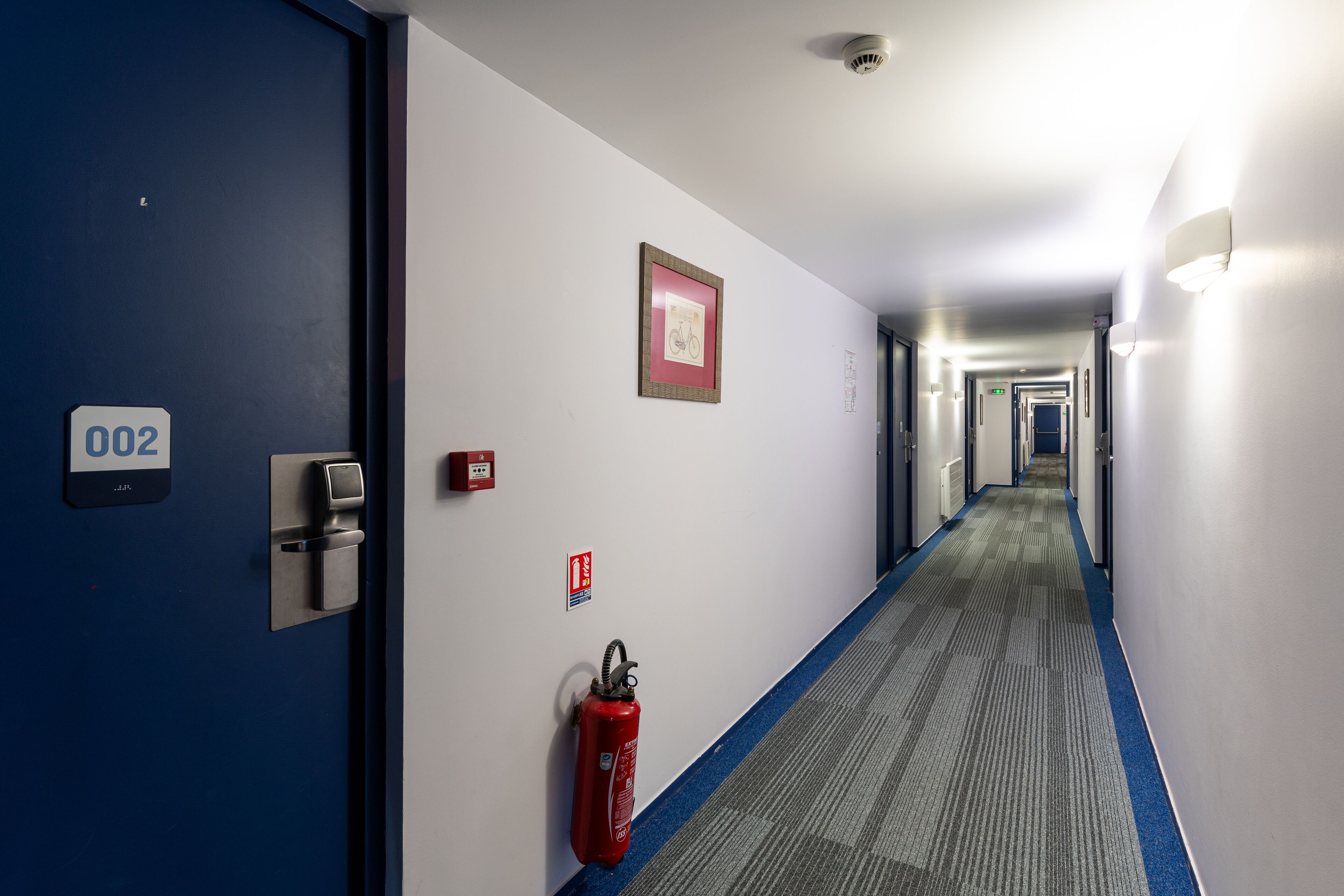 A hotel hallway with a blue door labeled '002', a fire extinguisher, and a wheelchair accessibility sign.
