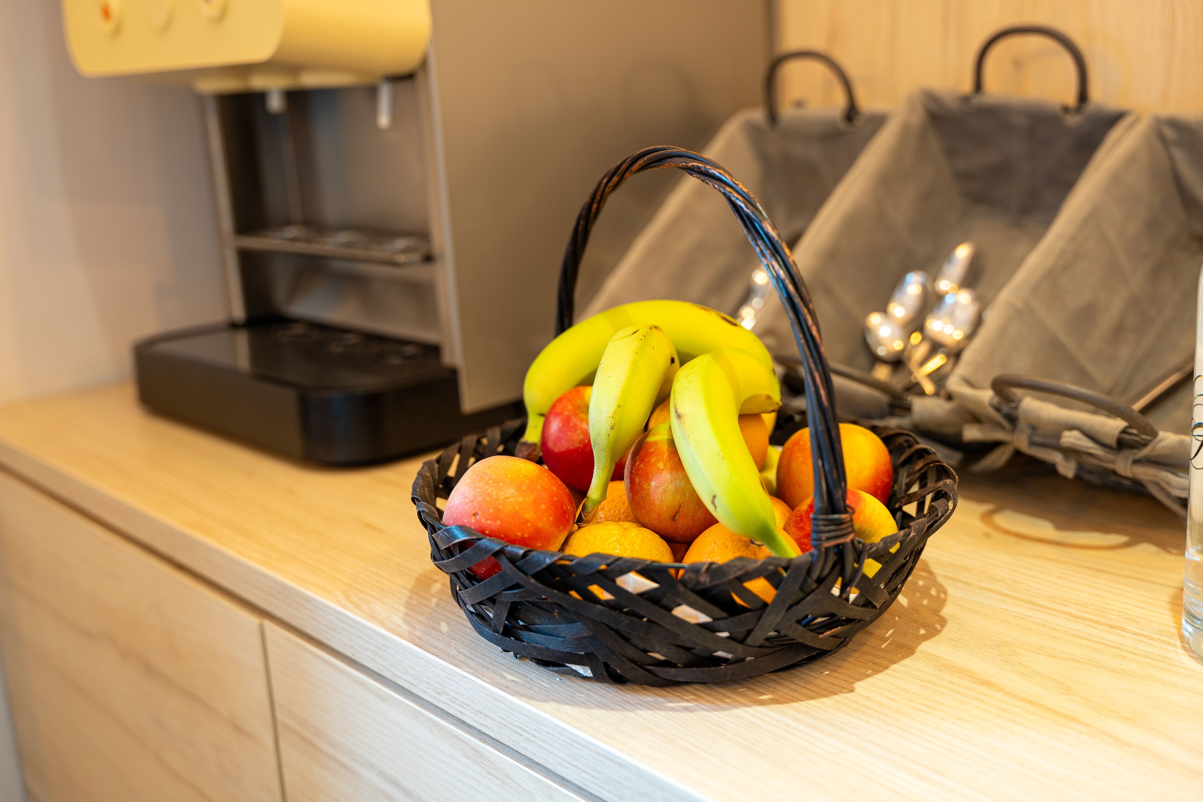 A basket of fruit on a kitchen counter with a coffee maker and bags in the background.