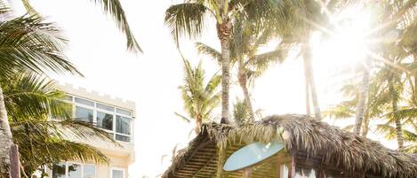 Una zona de playa con escalones que conducen a una estructura de techo de paja, palmeras y un edificio al fondo.