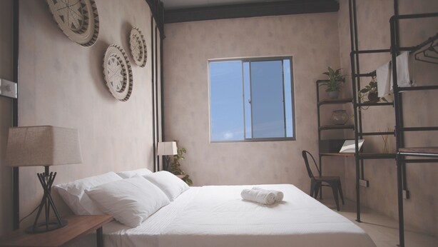 Une chambre à coucher avec un lit, une table de chevet, une lampe, une fenêtre et une étagère avec des plantes.