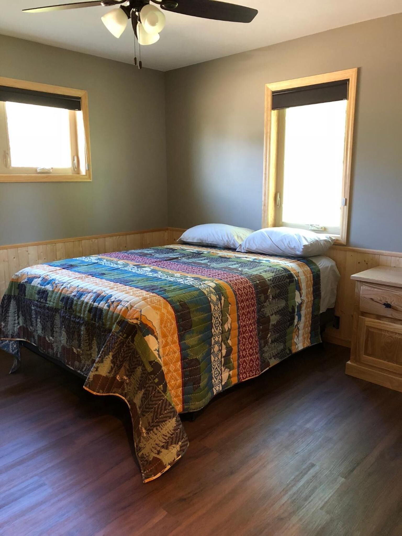 Quarto com cama, mesa de cabeceira de madeira, duas janelas e um ventilador de teto.