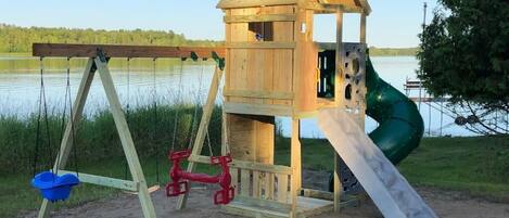 A wooden play structure with a slide and swings by a lake.