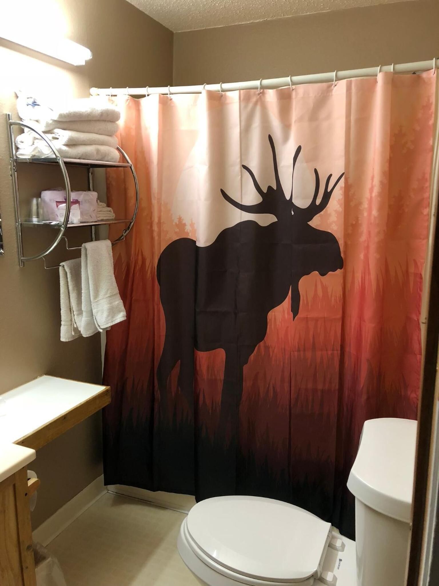 A bathroom with a shower curtain featuring a moose silhouette, a toilet, a sink with a towel rack, and a shelf with toiletries.
