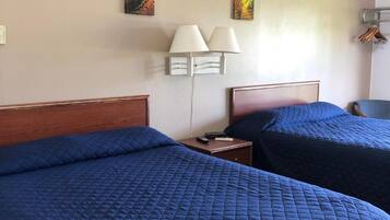 A hotel room with two beds, each with a blue quilted bedspread. There are three wall-mounted paintings depicting autumn foliage. A white lamp with two shades is on a bedside table.