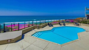 A pool with a view of the ocean, surrounded by a deck with lounge chairs and a table.