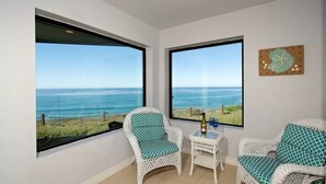 A room with two large windows offering a view of the sea, a white wicker chair with a green cushion, a small white table with a bottle, and a wall-mounted decorative piece.
