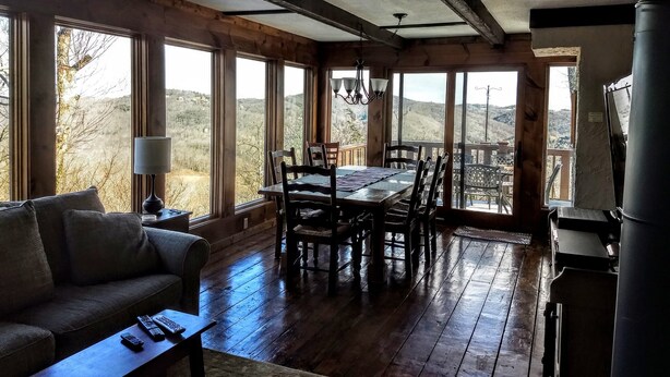 A spacious room with a wooden floor, large windows offering a view of the outdoors, a dining table with chairs, and a sofa. - Rustic farmhouse on Appalachian  Ski Mountain (Blowing Rock)