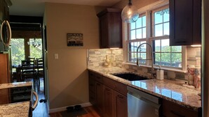 A kitchen with dark wooden cabinets, a granite countertop, a stainless steel dishwasher, and a window with a view of greenery. - Rustic farmhouse on Appalachian  Ski Mountain (Blowing Rock)