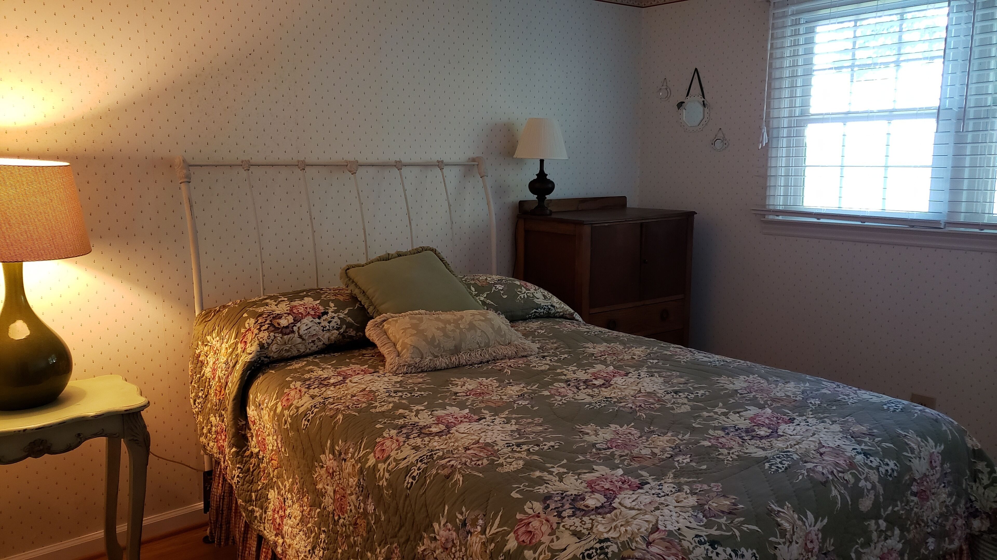 A bedroom with a floral bedspread, a bedside lamp, a wooden nightstand, and a window with blinds.
