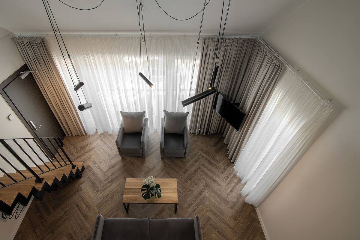 Un salon moderne avec un parquet en bois à motifs chevrons, un canapé, deux fauteuils, une table basse agrémentée d’une plante, et un escalier avec des rampes noires.