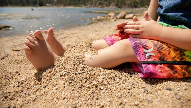 Un enfant est assis sur une plage de sable, avec un lac et une forêt en arrière-plan.