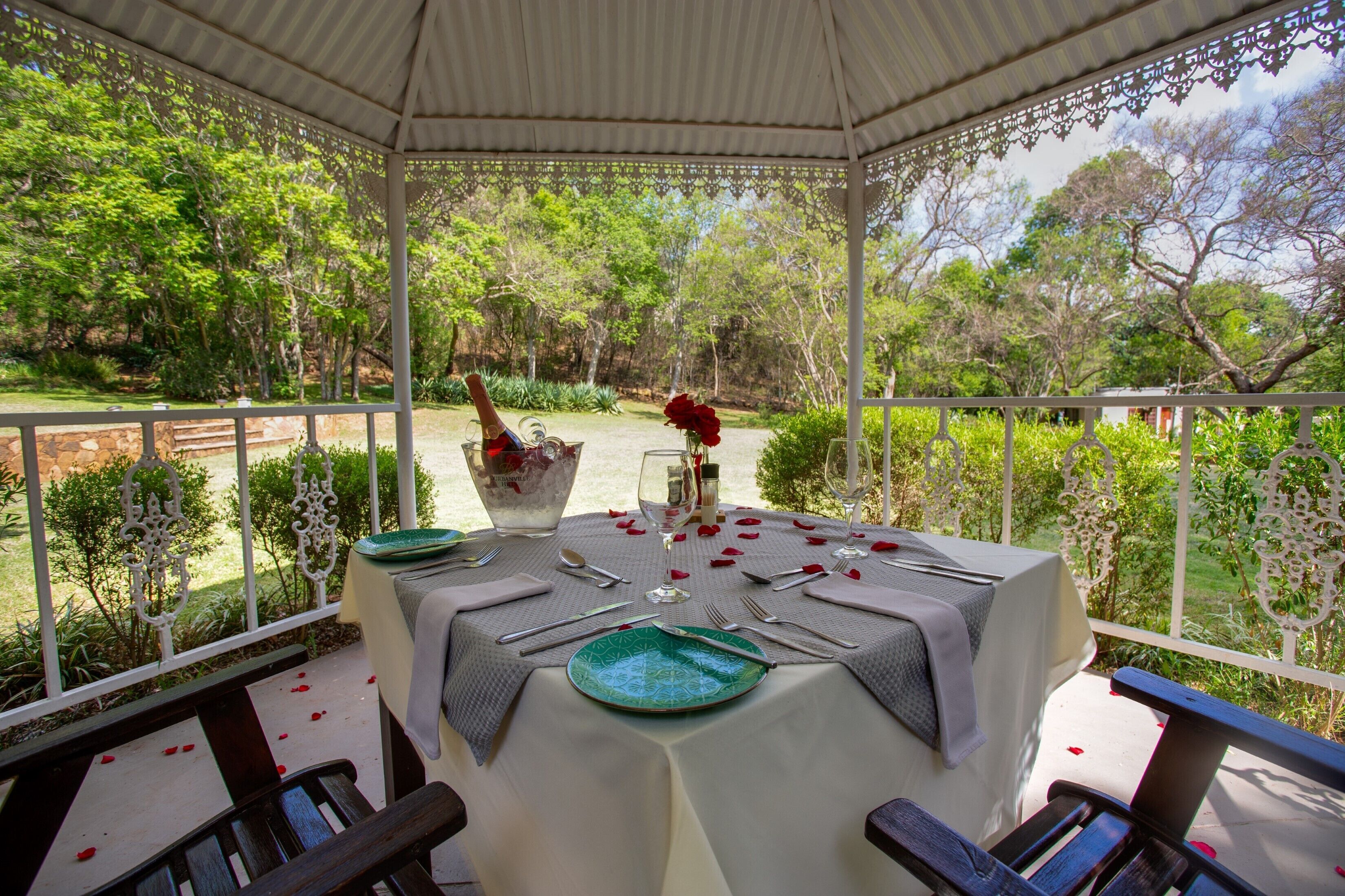 An outdoor dining area with a table set for two, surrounded by greenery and a gazebo structure.