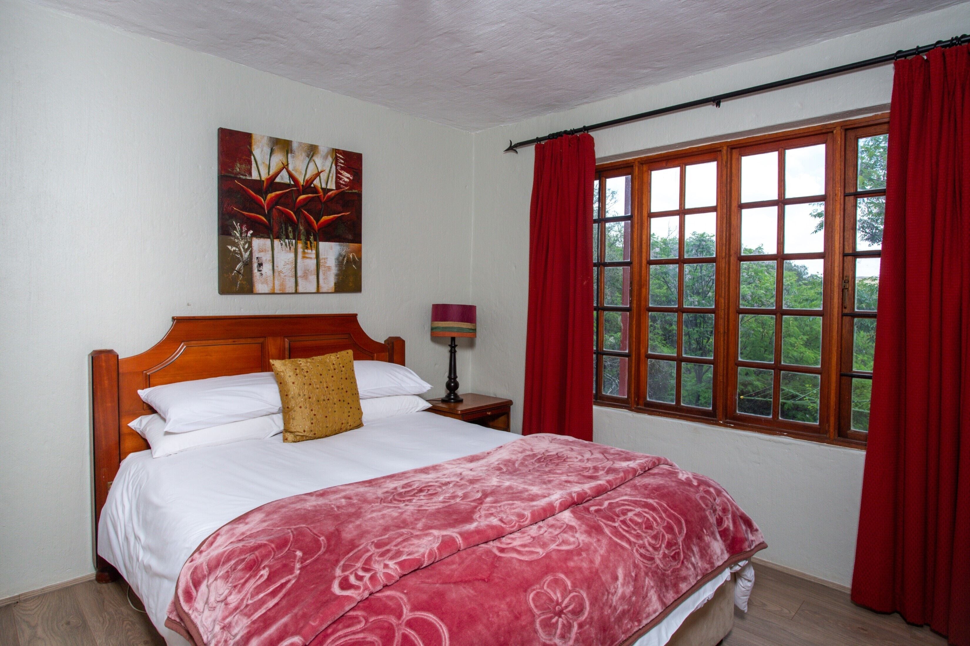 A bedroom with a wooden bed, a red curtain, a window with a view of trees, and a painting on the wall.
