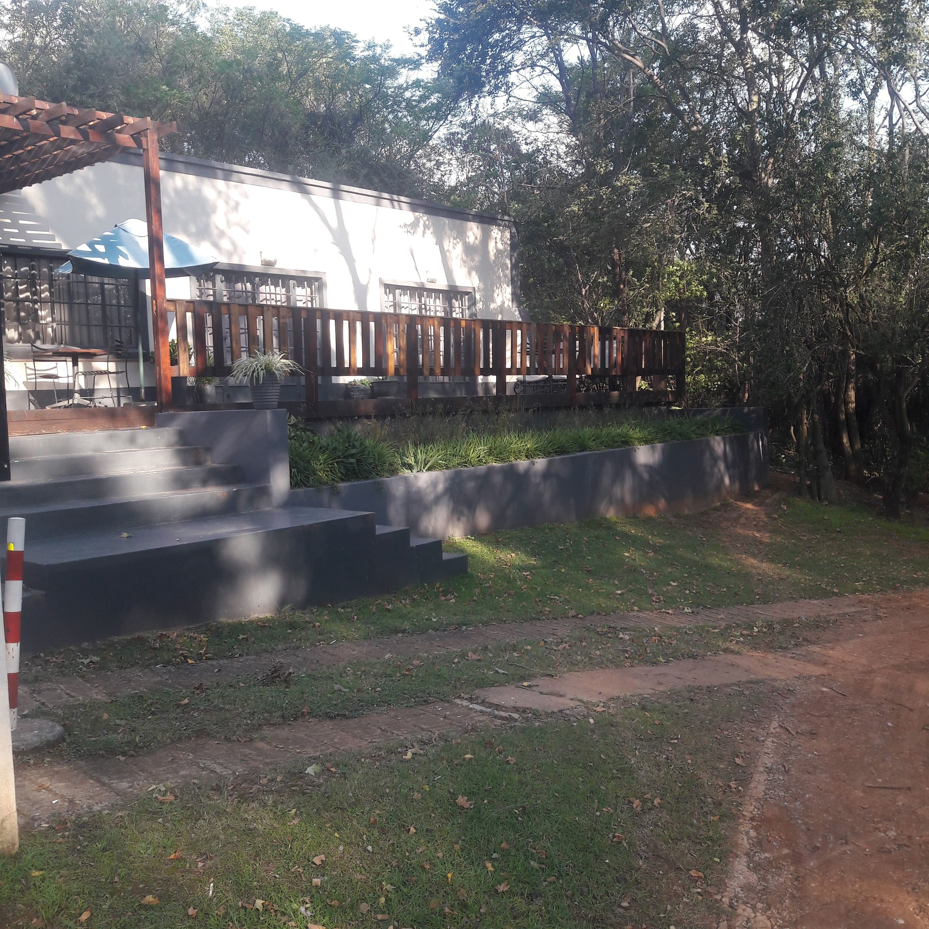 A building with a covered outdoor seating area, steps leading to a terrace, and a surrounding garden with trees.