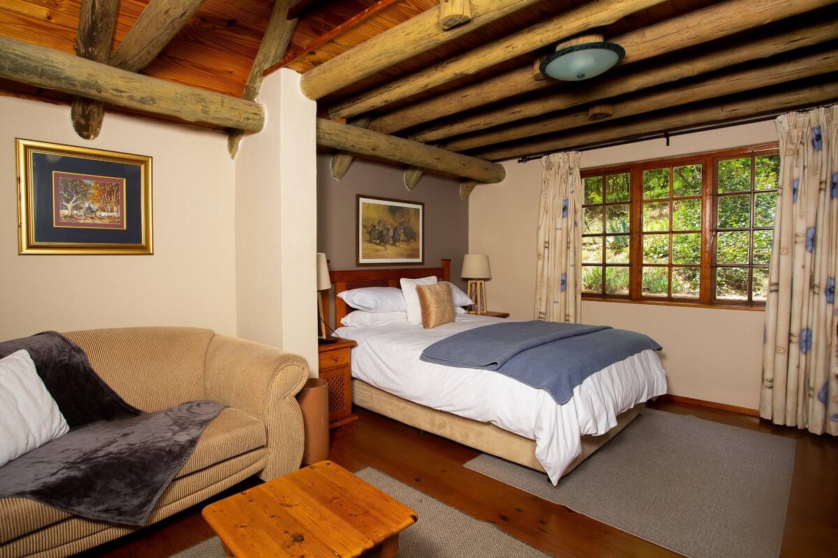 Ein Schlafzimmer mit Holzboden, einem Bett mit weißer Bettwäsche, einem Holztisch, einer Couch und einem Fenster mit Vorhängen.