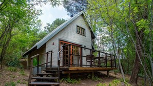 A rustic cabin with a wooden deck, a brick pathway leading to the entrance, and surrounded by dense forest. - Whispering Pines Country Estate (Magaliesberg)