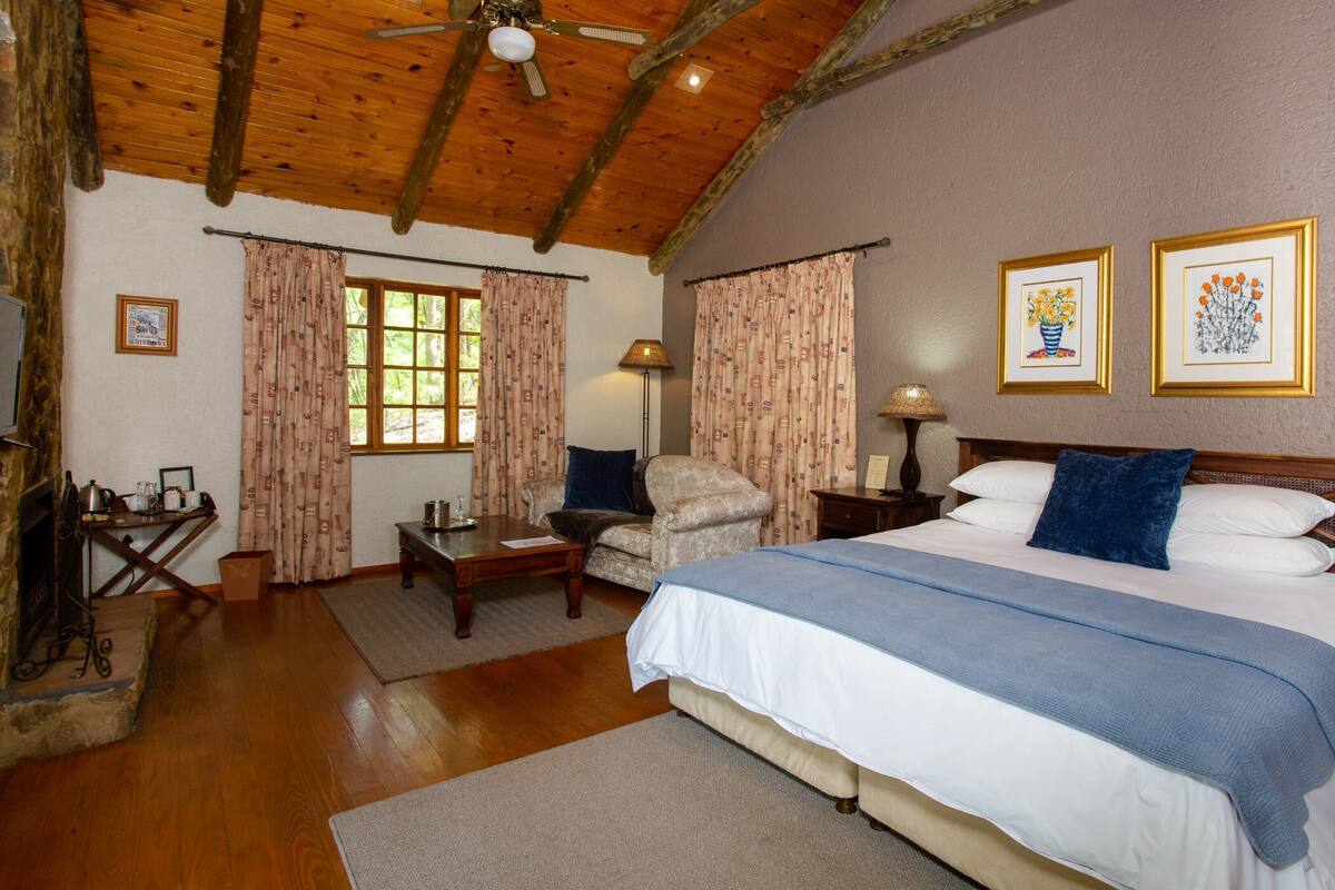 Ein gemütliches Schlafzimmer mit Holzbalkendecke, einem großen Bett mit blauer Decke, einem Sessel, einem Nachttisch und zwei gerahmten Bildern an der Wand.