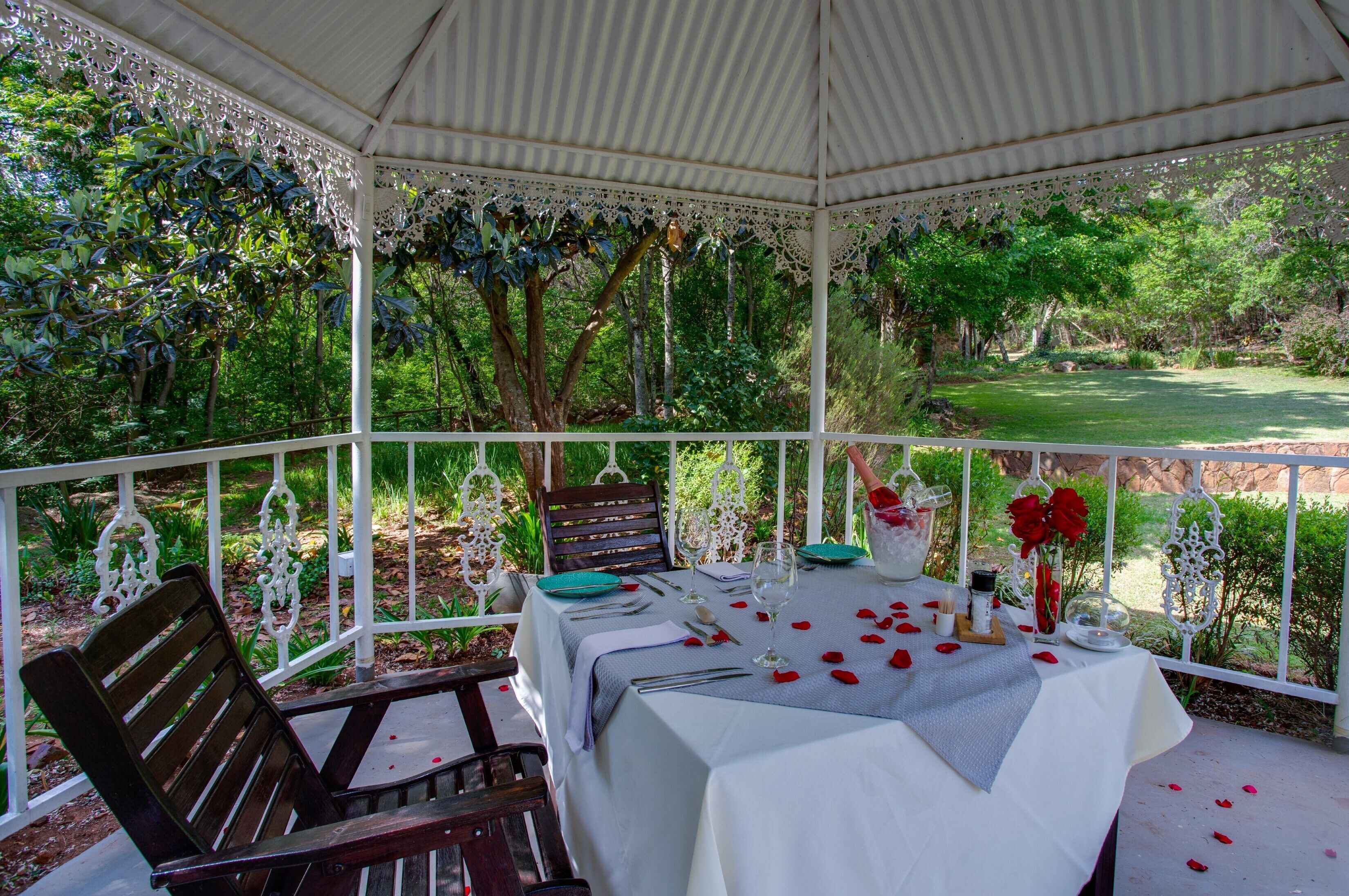 Un espacio cubierto para comer al aire libre con una mesa dispuesta para dos personas, rodeado de barandillas blancas y abundante vegetación.