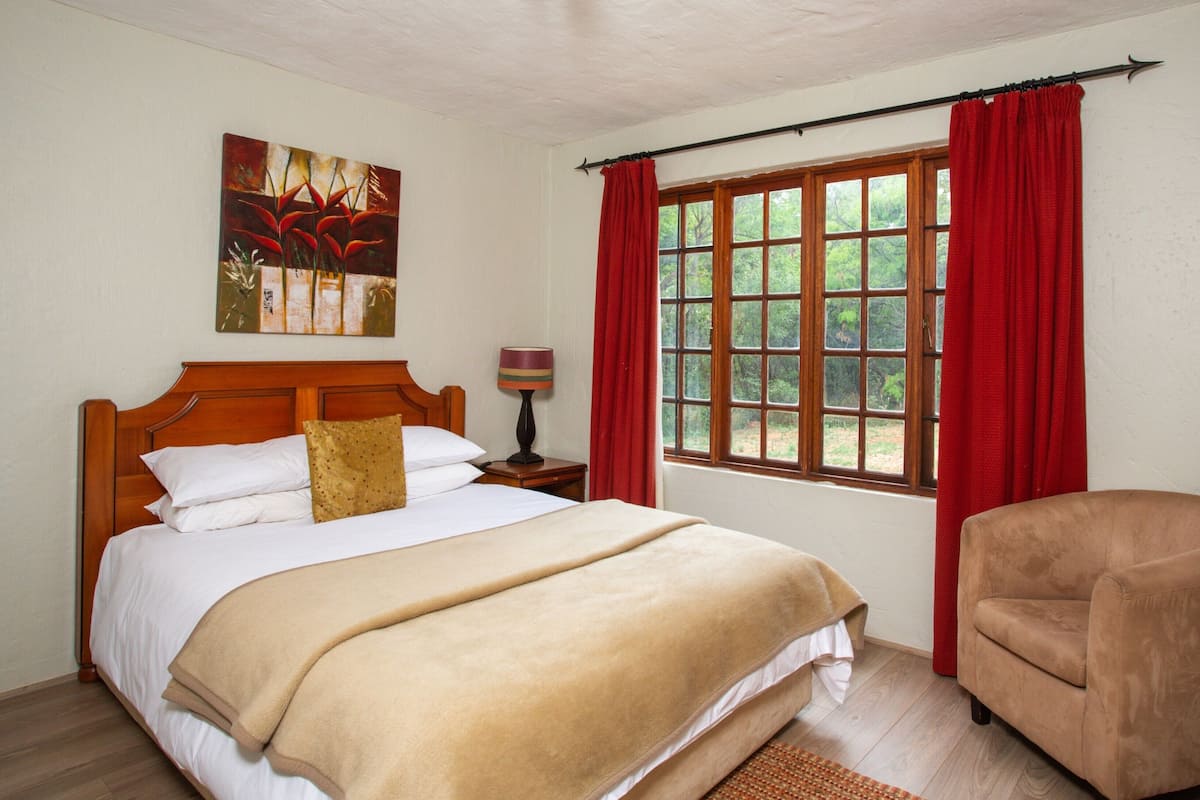 Ein Schlafzimmer mit einem großen Bett, einem hölzernen Kopfteil, einem Nachttisch mit Lampe, einem Sessel und einem Fenster mit roten Vorhängen.