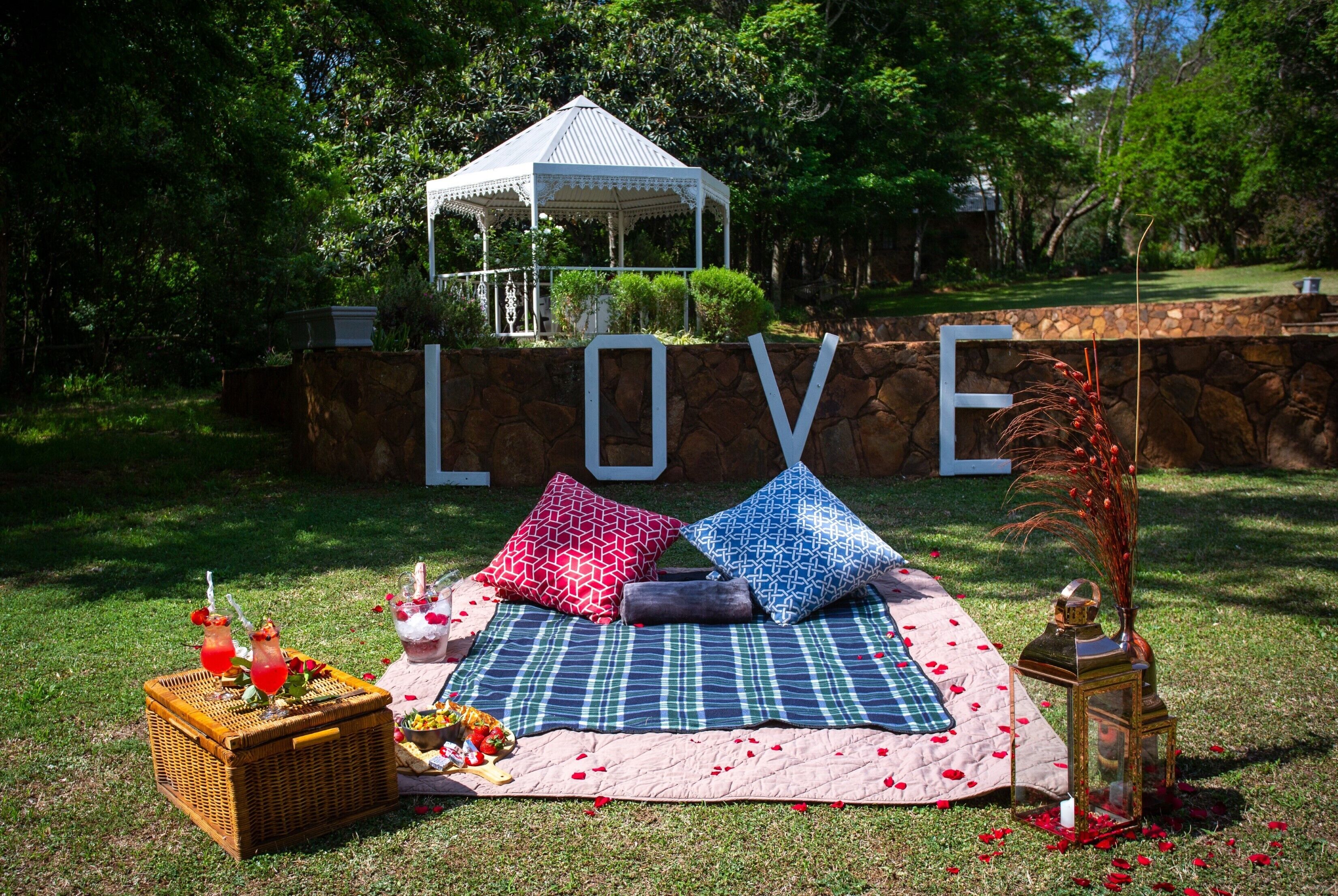 A picnic setup with a blanket, pillows, a basket, and a lantern, set against a backdrop with a large 'LOVE' sign and a gazebo.