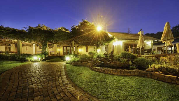 A well-lit garden pathway leading to a building with a stone facade, outdoor seating, and a small pond. - Whispering Pines Country Estate (Magaliesberg)