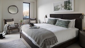 Un dormitorio con una cama grande, una silla, una mesita, una lámpara y una ventana.