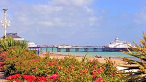 A coastal scene with a pier extending into the sea, a building with domed architecture, and a beach with people and flowers in the foreground. - Beachside Apartment (Eastbourne)