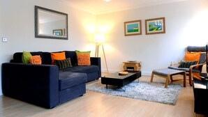 A living room with a dark blue sofa, a small wooden coffee table, a grey rug, a floor lamp, and a chair with a cushion. - Beachside Apartment (Eastbourne)