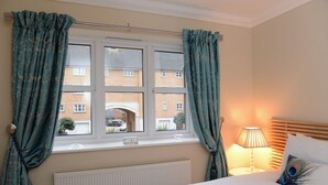 A bedroom with a bed, a window with curtains, a radiator, a bedside lamp, and a peacock pillow. - Beachside Apartment (Eastbourne)
