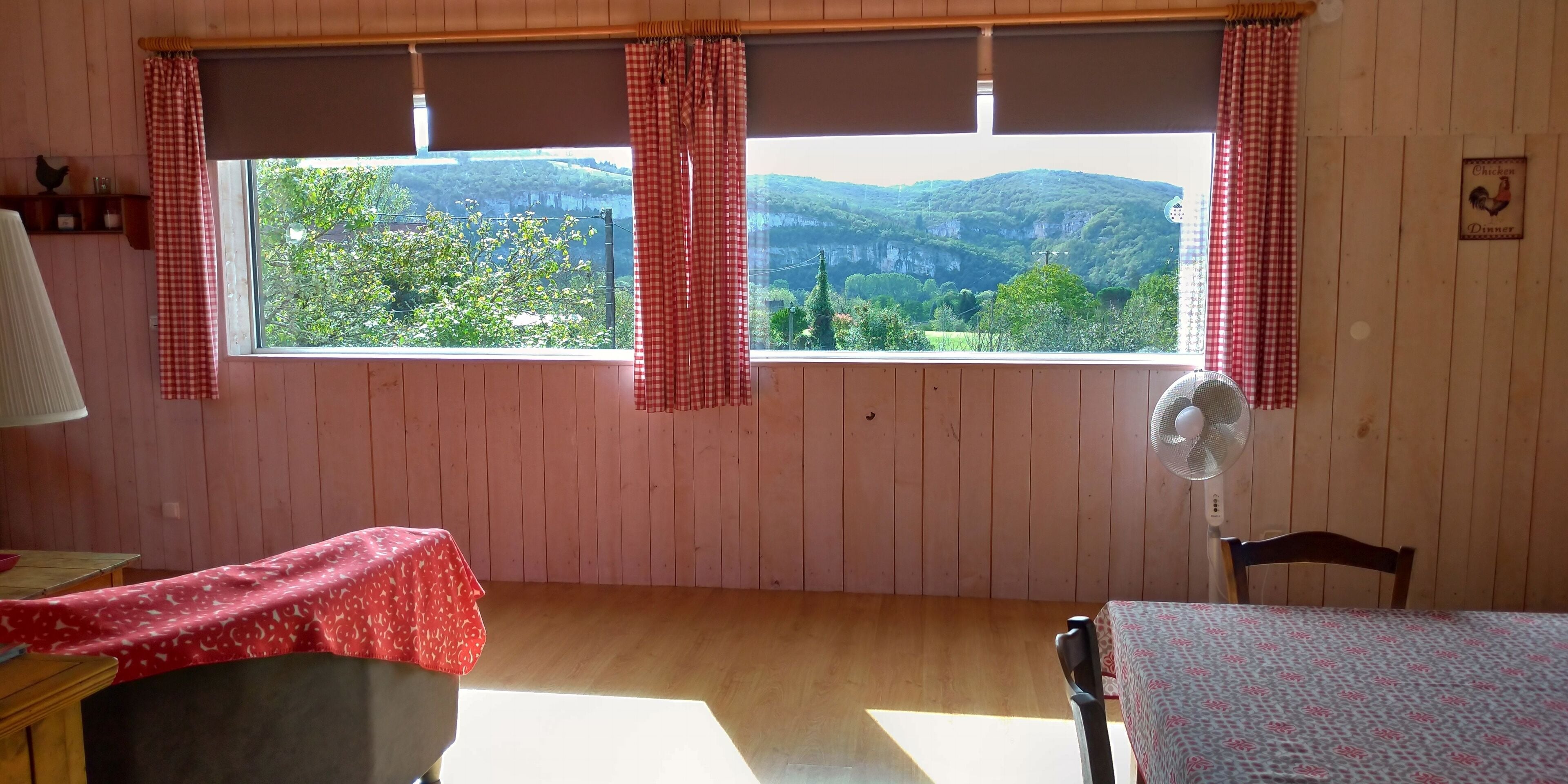 Una habitación con paredes de madera, un ventanal con cortinas a cuadros rojos y blancos, un ventilador y una mesa con mantel rojo.