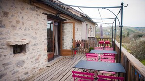 Un edificio de piedra rústico con una terraza de madera, asientos exteriores con sillas rosas y vistas al campo.