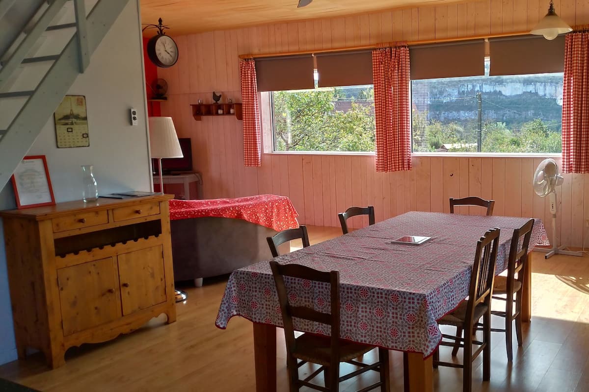 Un rincón comedor acogedor con una mesa dispuesta para cuatro, sillas de madera, un aparador de madera, una persiana de ventana a cuadros rojos y blancos, y un reloj en la pared.