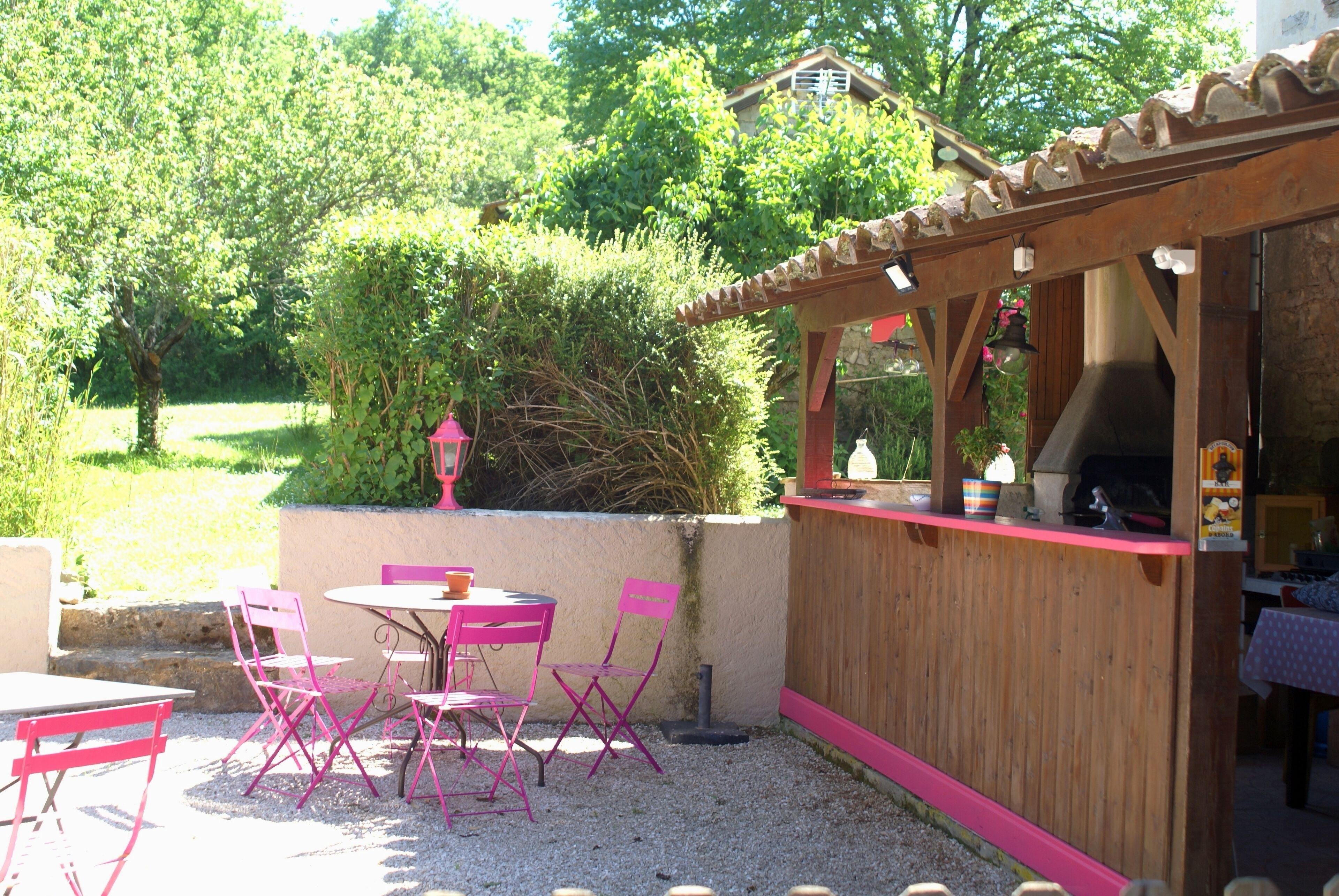 Un patio al aire libre con zona de barbacoa de madera, rodeado de grava y sillas rosadas.