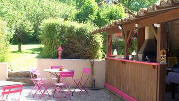 Un patio al aire libre con zona de barbacoa de madera, rodeado de grava y sillas rosadas.