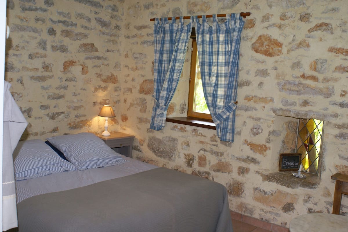 Un dormitorio con pared de piedra, cama con ropa de cama a rayas, mesita de noche con lámpara y ventana con cortinas escocesas.