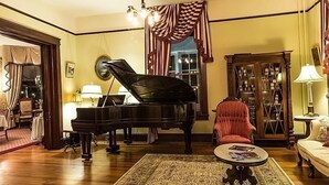 A grand piano in a well-lit living room with a patterned rug, a red armchair, and a bookshelf. - Double Oaks Bed and Breakfast (Greensboro)