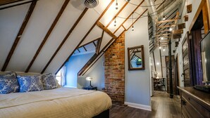 A bedroom with a sloped ceiling, exposed wooden beams, a brick wall, and a large bed with patterned pillows. - Double Oaks Bed and Breakfast (Greensboro)