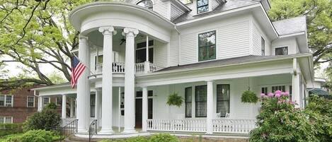A large, two-story house with a wrap-around porch, a flag, and a well-maintained garden.