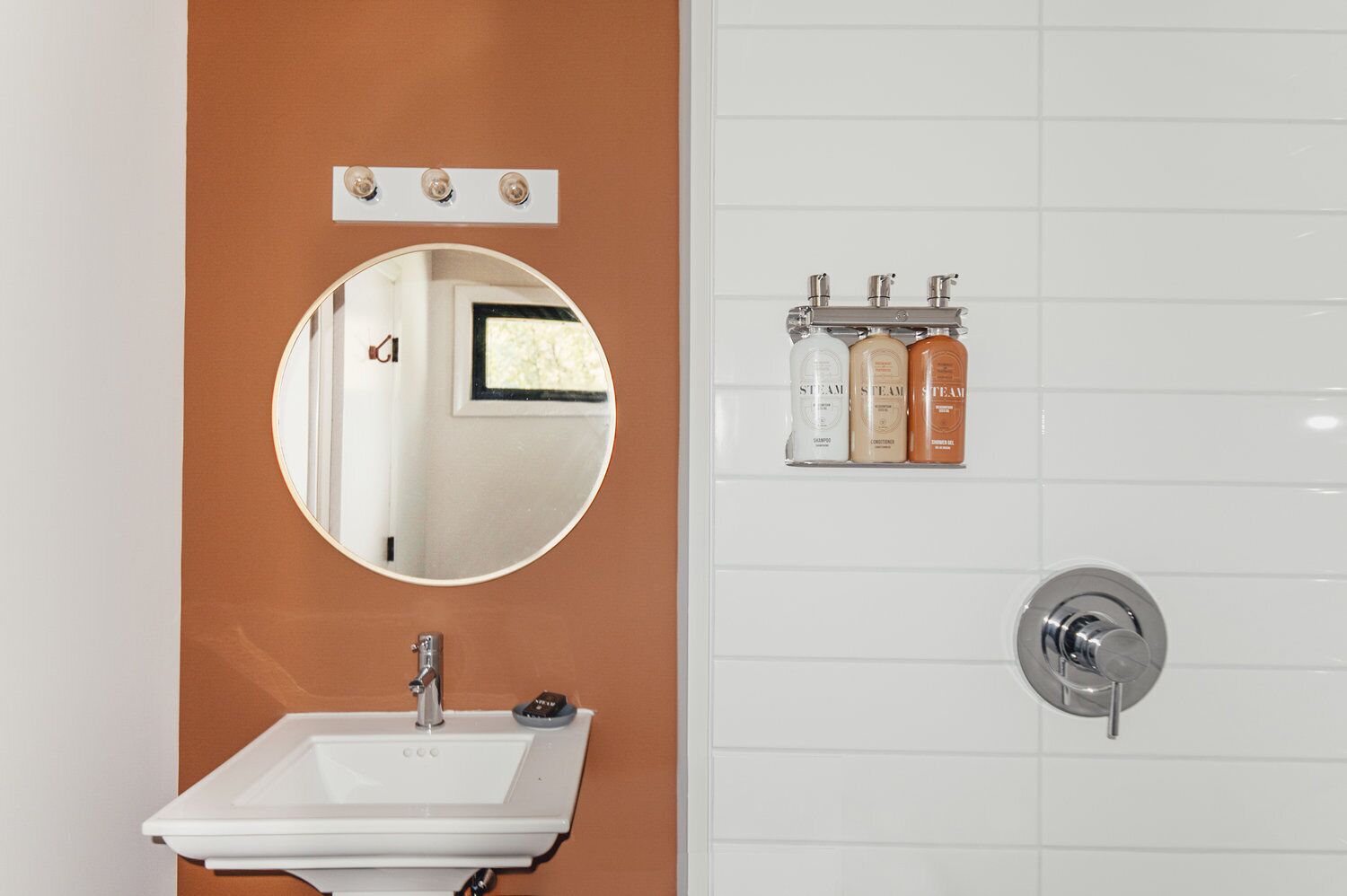 Un bagno con un lavandino bianco, uno specchio rotondo e un water. In parete, tre flaconi di shampoo e balsamo.