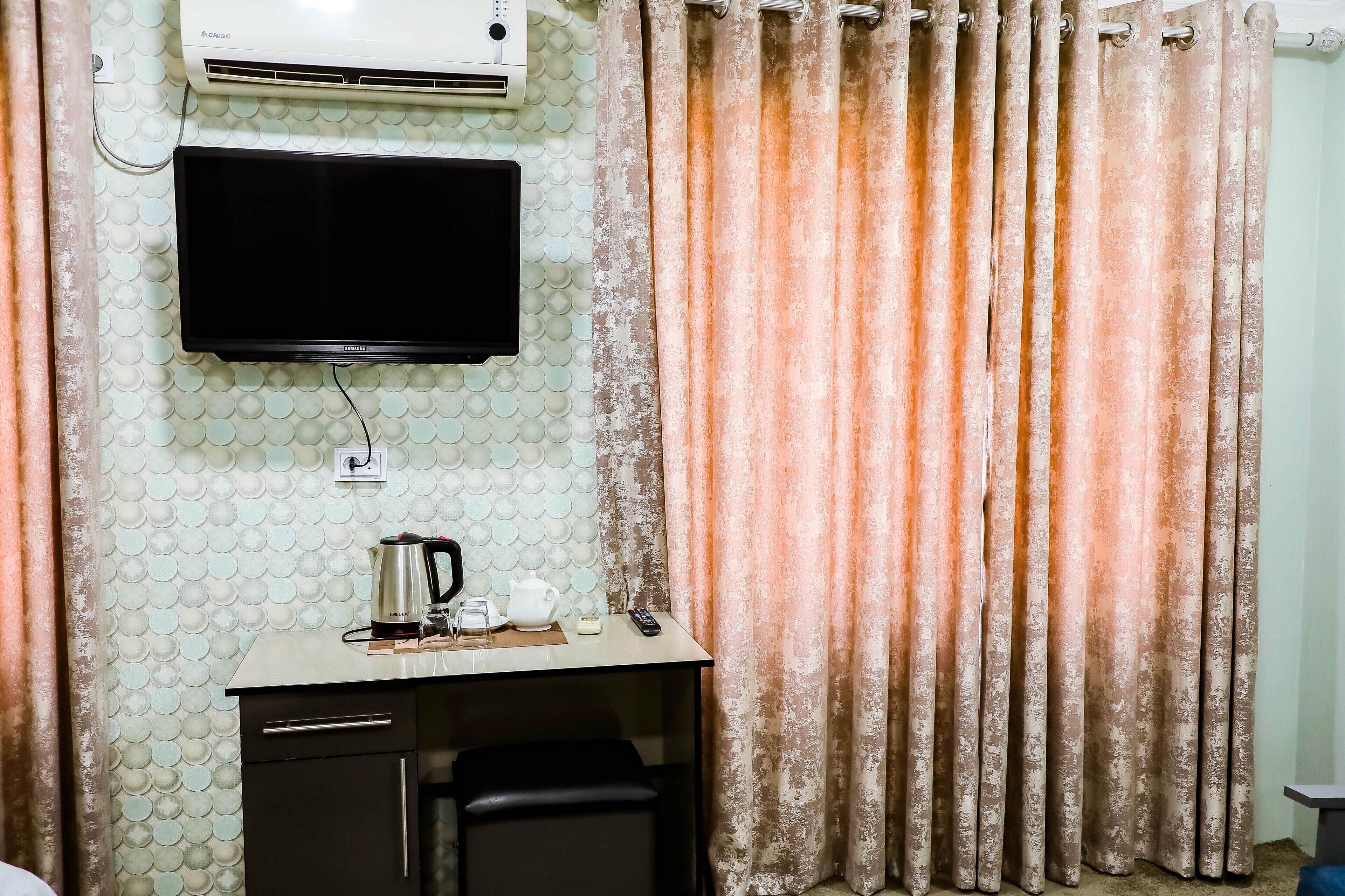 A room with a flat-screen TV mounted on the wall, a small table with a kettle, a remote control, and a candle. There is a curtain with a textured pattern, and a wall-mounted air conditioner.