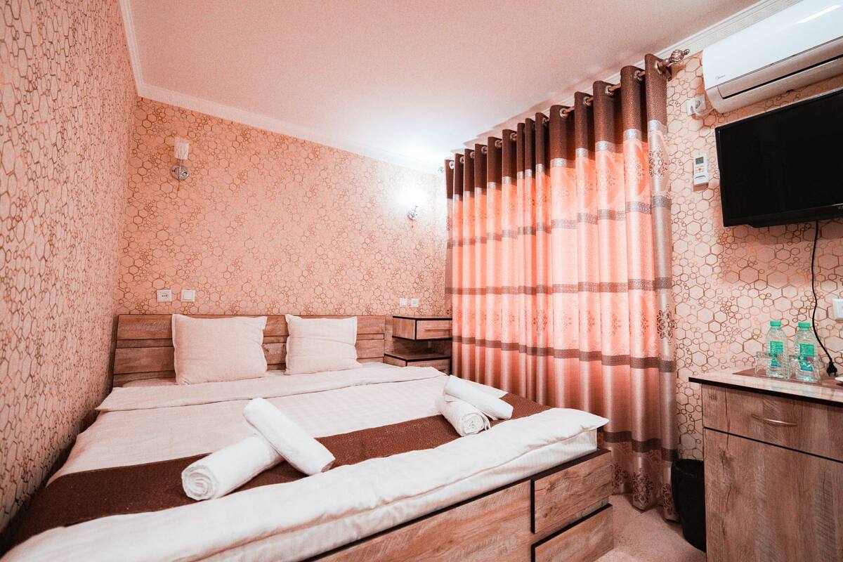A hotel room with a bed, wooden headboard, two bedside tables, a TV, and a window with curtains.