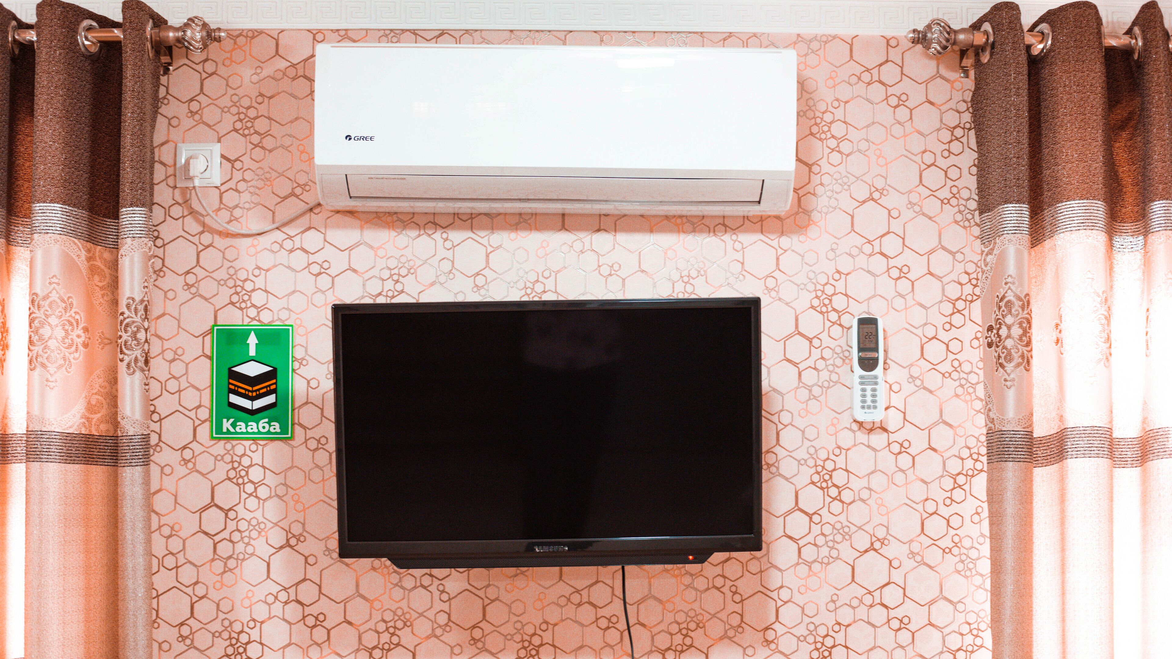 A flat-screen TV mounted on a wall with a patterned wallpaper, a thermostat, and a remote control on the wall.
