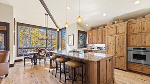 A modern kitchen with wooden cabinets, a large island with stools, and a view of autumn trees through large windows. - Grand Hyatt|Bright & Airy Centrally Located Condo (Vail)