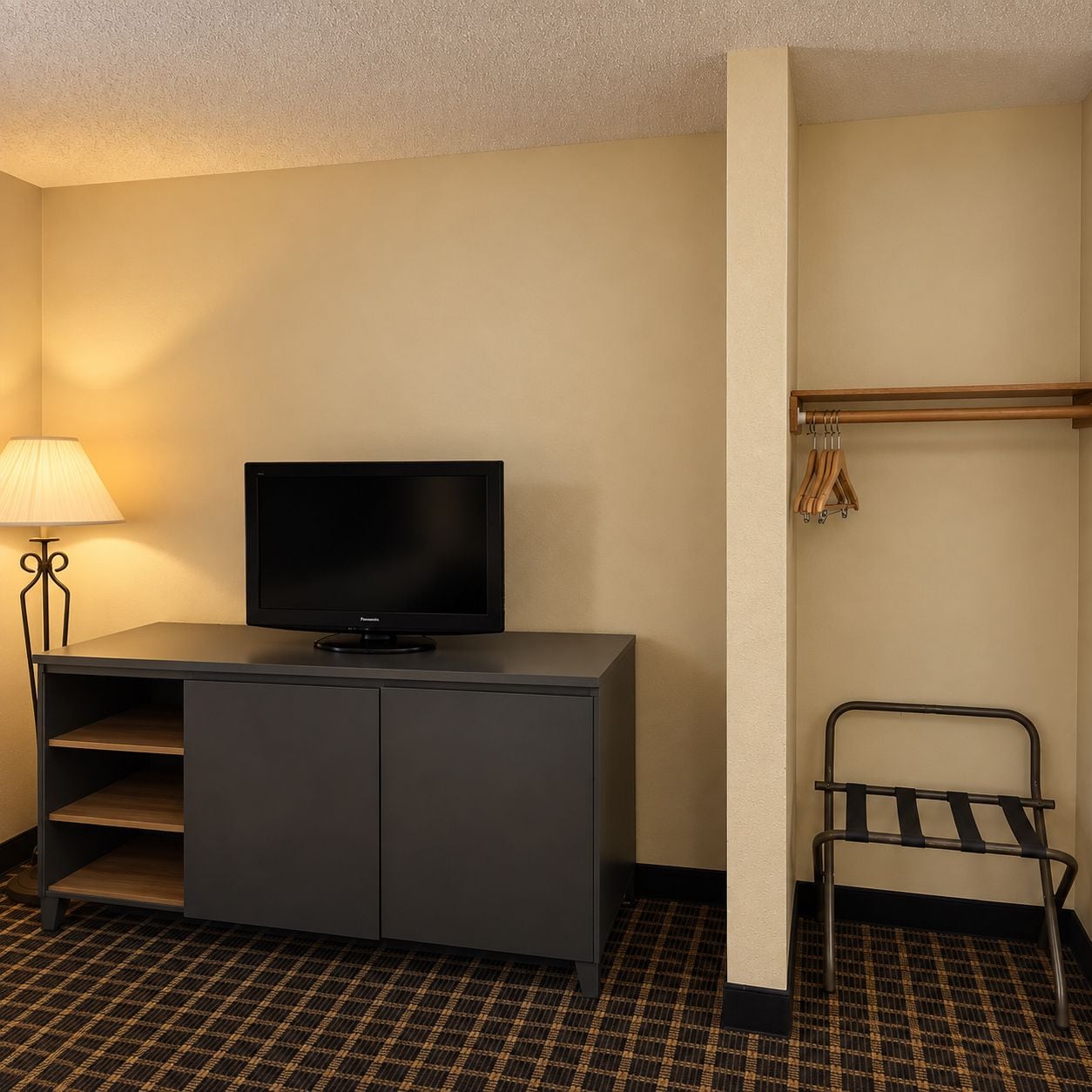 A hotel room with a flat-screen TV on a dark-colored cabinet, a lamp on the left, and a coat rack with hangers to the right.