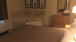 Habitación de hotel con una cama grande, una mesita de noche con lámpara, un aparador, un espejo y un cuadro en la pared.