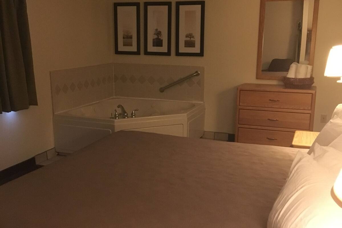 Ein Hotelzimmer mit einem großen Bett, einem Nachttisch mit Lampe, einem Schminktisch, einem Spiegel und einem gerahmten Bild an der Wand.